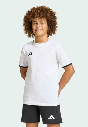 Ragazzo con capelli ricci che indossa una maglietta bianca Adidas e pantaloncini neri Adidas, in piedi con le mani dietro la schiena su sfondo chiaro.