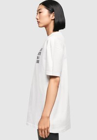 Weißes Baumwoll-T-Shirt mit kurzen Ärmeln, oversized Schnitt und schwarzem Aufdruck auf der Vorderseite. Einheitliche Farbe, glatte Textur, Rundhalsausschnitt.