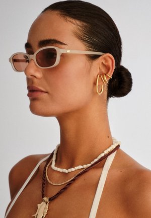 Woman with sleek bun wears rectangular beige sunglasses, three gold earrings on left ear, and layered white and brown beaded necklaces.