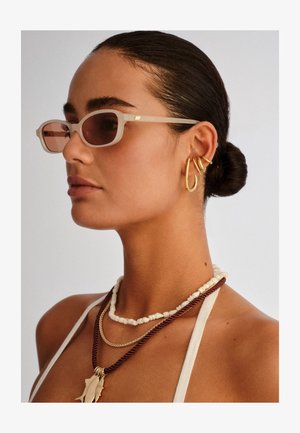 Γυναίκα με κομψό σινιόν φοράει ορθογώνια μπεζ γυαλιά ηλίου, τρία χρυσά σκουλαρίκια στο αριστερό αυτί και στρωμένα λευκά και καφέ κολιέ με χάντρες.