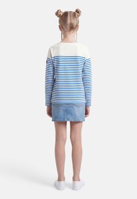 Παιδί με διπλούς κότσους, φορώντας λευκό μπλουζάκι με μπλε ρίγες μακρυμάνικο, φούστα ανοιχτού τζιν και λευκά αθλητικά παπούτσια, στέκεται γυρισμένο με την πλάτη.