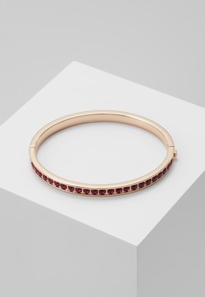 Kultainen ranneke, jossa on rivi pieniä pyöreitä punaisia jalokiviä ulkoreunalla, esillä valkoisella pinnalla.