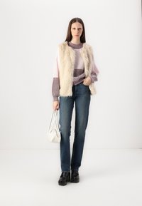 Gilet sans manches en fausse fourrure sur un pull rayé rose et bordeaux, jeans bleus et chaussures à plateforme noires; tenant un sac à main blanc.