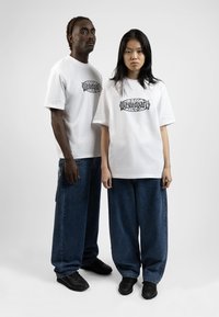 Two individuals wearing white graphic t-shirts with a black logo, paired with loose-fitting blue denim jeans and black shoes.