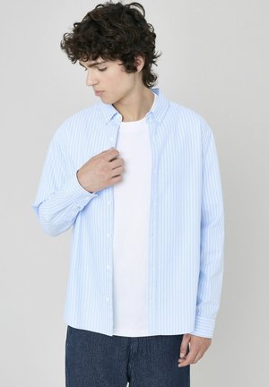Chemise boutonnée rayée bleu clair en coton, avec un col, des manches longues et une coupe décontractée sur un fond neutre.