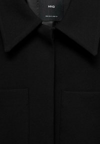 Veste noire à col en tissu texturé, avec deux poches avant et une étiquette au niveau du col. Design simple et élégant.