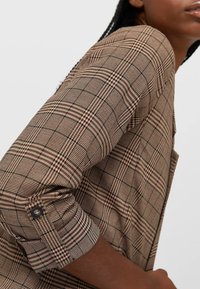 Hnědá kostkovaná košile s klasickým vzorem, zapínacími rukávy a hladkou texturou látky. Design zahrnuje tenké linie v kontrastních barvách.