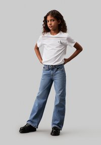 T-shirt bianco con un piccolo logo nero, jeans blu a gamba larga e scarpe nere. Capelli ricci e lasciati sciolti. Outfit semplice e casual.