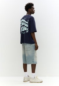 Navy oversized t-shirt featuring bold light blue text on the back. Paired with light denim Bermuda shorts and white sneakers.