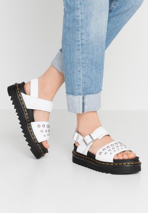 Fødder iført hvide sandaler med nitter og sorte tykke såler kombineret med opkørte lyseblå jeans mod en ensfarvet baggrund.