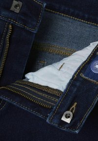 Denim azul oscuro con costuras doradas. Cuenta con un cierre de cremallera metálica y botón, además de un forro de tela azul claro visible en el interior.