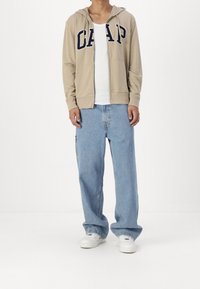 Sweat à capuche beige zippé avec logo "GAP", t-shirt blanc et jean bleu clair ample, porté avec des baskets blanches. Tissu lisse avec une coupe décontractée.