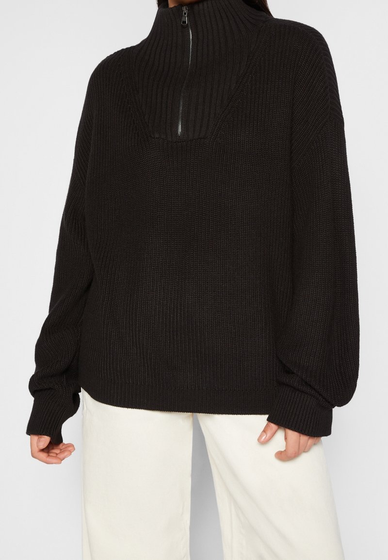 Personne portant un pull en maille côtelée noir avec un col à demi-fermeture éclair et des manches surdimensionnées, associée à un pantalon blanc.