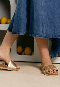 Deux pieds portant des sandales sur une surface beige texturée. Le pied gauche est dans une glissière brillante de couleur marron clair avec une boucle. Le pied droit porte une glissière de couleur beige.