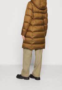 Abrigo acolchado marrón con capucha, con costuras horizontales y un diseño de longitud media. Se lleva con pantalones beiges claros y botas gruesas negras.