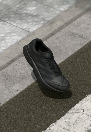 Czarny sportowy sneaker z sznurowadłami spoczywający na fakturowanej szarej nawierzchni w pobliżu podniesionej krawędzi chodnika i powierzchni z nawierzchnią dotykową.