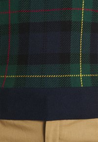 Suéter con patrón verde y azul marino con diseño a cuadros, con acentos rojos y amarillos. Dobladillo acanalado en azul marino. La tela parece suave y con textura.