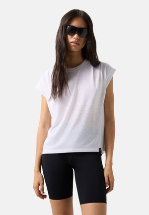 Femme portant de grandes lunettes de soleil noires, un haut blanc sans manches avec des épaules rembourrées et un short de cycliste noir, debout devant un fond uni.