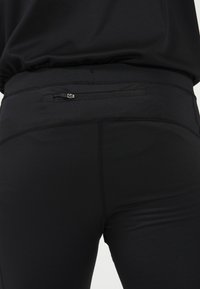 Svarta atletiska leggings med en mjuk textur, åtsittande design och en diskret dragkedjeficka på baksidan, med platta sömmar.