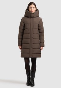 Cappotto puffo marrone con colletto alto, chiusura a bottoni e design imbottito. Presenta una tasca frontale ed è lungo fino al ginocchio. Indossato sopra leggings neri e stivali.