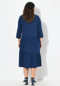 Vestito blu navy fino al ginocchio con maniche a 3/4, orlo a balze a strati e texture liscia. Indossato con sneaker alte blu.