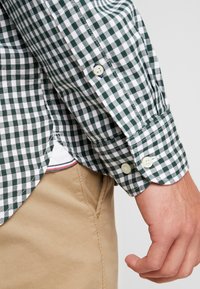 Camisa de cuadros verde y blanca con mangas largas, botones claros y detalles cosidos en el dobladillo, combinada con pantalones beige.