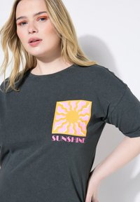 Camiseta gris de manga corta con un gráfico cuadrado que presenta un sol amarillo rodeado de rayos rosas y el texto "SUNSHINE" en rosa.