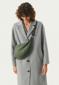 Borsa a tracolla in tessuto verde con forma arrotondata, dotata di una larga tracolla regolabile e una superficie liscia, indossata sopra un cappotto grigio.