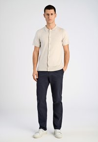 Beige kortärmad polo med knapplist, i kombination med marinblå byxor och vita sneakers, stående mot en ljus bakgrund.
