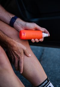 Main tenant un flacon vaporisateur MOVE orange contre une jambe pliée avec une montre de sport noire et une chaussette noire et blanche visibles.