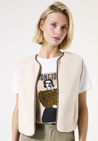 Chaleco de forro polar beige con detalles en marrón, que presenta un corte relajado. Lleva encima una camiseta blanca con un diseño gráfico de estampado de leopardo y una ilustración negra.