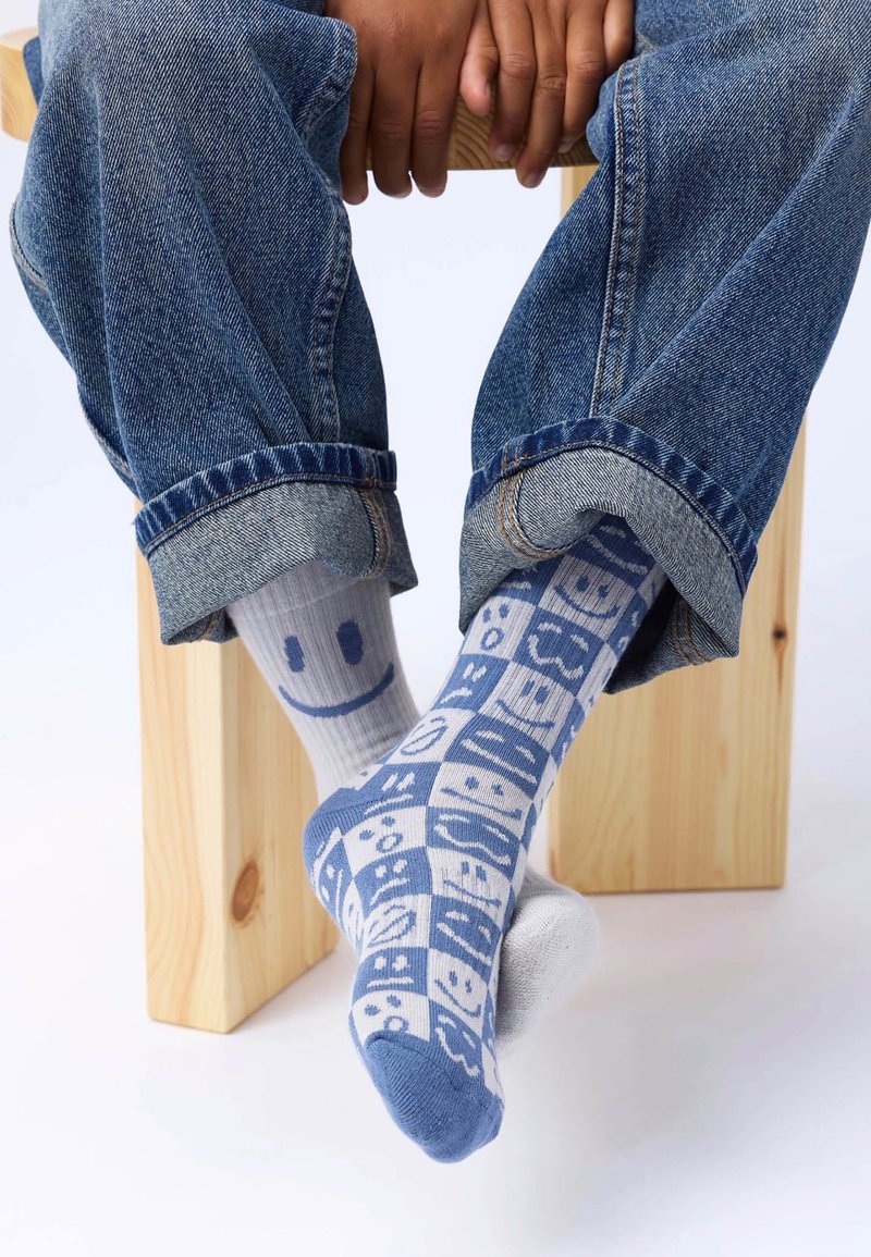 Fargerike sokker med et mønster av smilende ansikter i blått og grått, kombinert med denimjeans, hvilende på en treblokk.