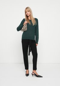 Mørkegrønn strikkegenser med bred utskjæring, svarte skreddersydde bukser med knyteliv, og svarte spisse slingback-heler. Mønstret skulderveske.