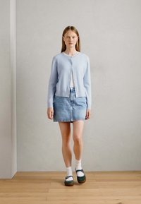 Ljust blå stickad kofta, vit t-shirt, denimkjol och gröna-bruna platåskor med vita strumpor. Rakt, minimalistisk design.