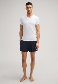 T-shirt blanc à manches courtes en V associé à un short de sport bleu marine foncé. Le modèle se tient pieds nus, les mains dans les poches, sur un fond neutre.