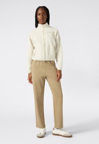 Giacca zip-up crema con colletto alto, abbinata a pantaloni beige e sneakers bianche. Tessuto liscio, design minimalista e tasche laterali.