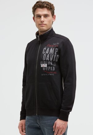 Junger Mann trägt eine schwarze Zip-Up-Jacke mit dem Text "CAMP DAVID RIDING THE WORLD" und steht vor einem schlichten hellen Hintergrund.
