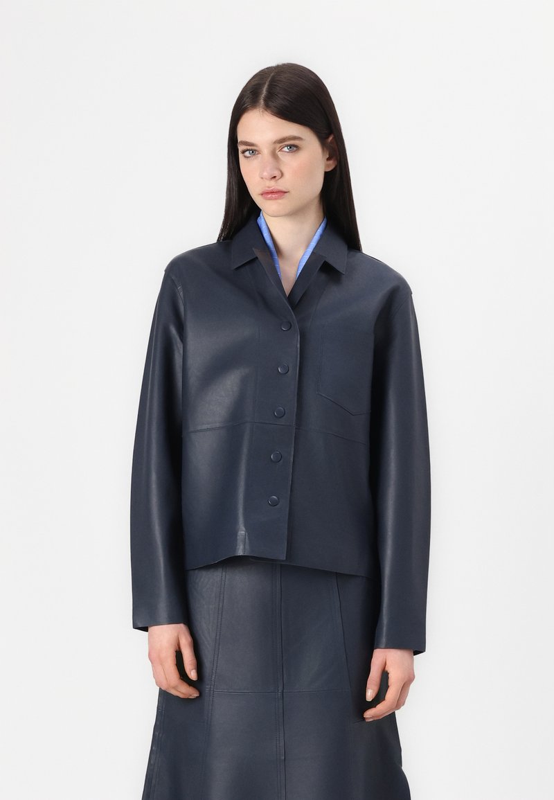Navy leather cropped jacket with a button-up front, pointed collar, and single chest pocket; paired with a matching leather skirt.