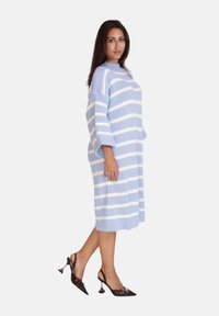 Strickkleid mit hellblauen und weißen vertikalen Streifen, knielang, mit weiten Ärmeln und Taschen vorne, kombiniert mit schwarzen hohen Schuhen.