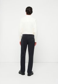 Weißes Langarmhemd kombiniert mit dunklen Marineblauen Hosen, die eine glatte Textur und unauffällige Gesäßtaschen aufweisen. Schwarze Schuhe vervollständigen den Look.