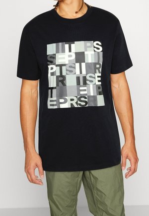 Mann trägt ein schwarzes T-Shirt mit einem grauen und weißen abstrakten Blockbuchstabenmuster und grüner Hose.
