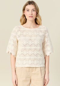 Top in pizzo color crema con maniche scallop e motivo a diamante, caratterizzato da una trama a maglia aperta e un colletto rotondo. Abbinato a pantaloni beige.