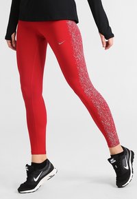 Czerwone legginsy sportowe z teksturowanym wzorem po jednej stronie, wykonane z elastycznego materiału. Połączone z czarnymi butami sportowymi z białym logo.