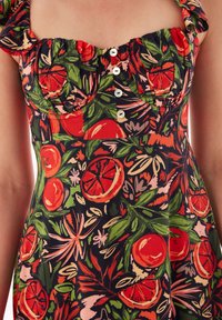 Blumenkleid mit schwarzem Hintergrund, das lebendige rot und grün gemusterte Früchte und Blätter zeigt. Akzentuiert mit weißen Knöpfen und gerafften Trägern.