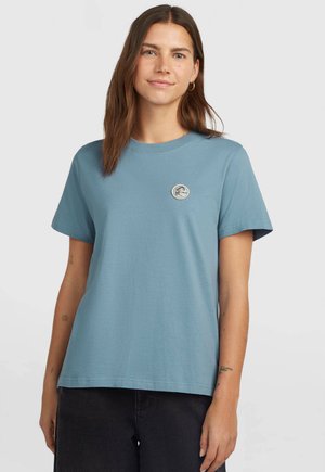 Jonge vrouw met lang bruin haar die een blauw T-shirt met korte mouwen draagt met een klein rond logo op de borst en een zwarte broek.
