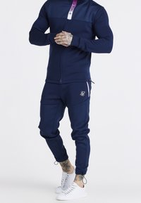 Tuta con zip color navy realizzata in tessuto morbido, con colletto accentato di viola, logo ricamato e sneakers bianche con lacci texturizzati.