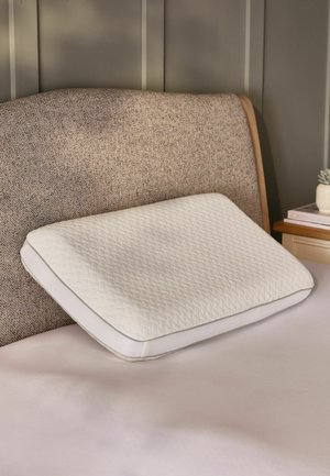 Weißes, strukturiertes Kissen, das auf einem beige bezogenen Kopfteilbett ruht, mit einem Beistelltisch, auf dem Bücher und eine kleine Pflanze in einem neutralen Raum stehen.