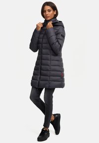 Padded lång kappa i mörkgrå med en figurnära design, huva, dragkedja framtill och subtilt varumärkesmärke. Matchad med mörka skinny jeans och sneakers.