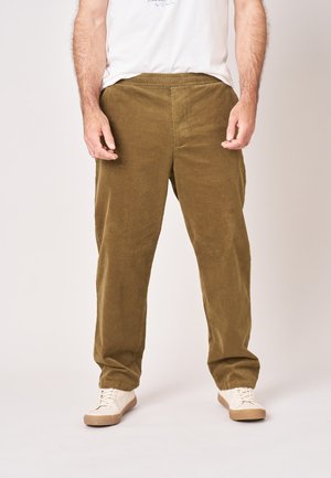 Man draagt olijfgroene corduroy broek, witte sneakers en een wit T-shirt, staand tegen een effen achtergrond.