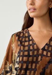 Blusa marrone con motivo, caratterizzata da un design a scollo a V, con un mix di griglie e forme astratte in toni terrosi e tessuto liscio e leggero.